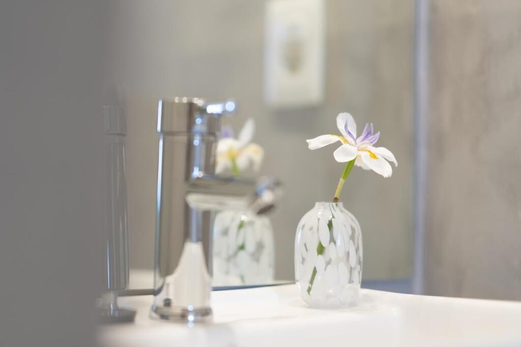 Une salle de bain avec un lavabo blanc, un vase en verre contenant une fleur et un miroir qui reflète le lavabo et le robinet.