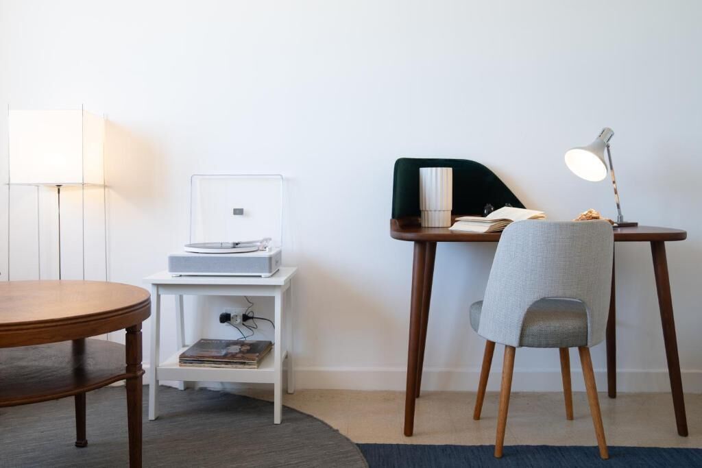 Una habitación con una mesa redonda de madera, una mesita auxiliar blanca con una lámpara, una silla y un escritorio con una lámpara y un libro.