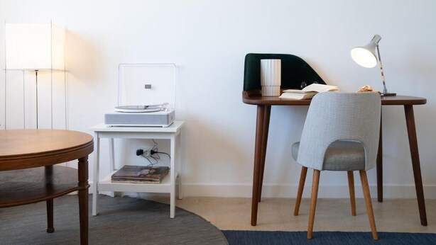 A room with a round wooden table, a white side table with a lamp, a chair, and a desk with a lamp and a book. - Vitaminas - Casa de Playa y Cafe (Punta del Este)