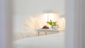 Una cama bien hecha con cabecera blanca, una mesita de noche con lámpara, una planta en maceta y un plato con un pastel.