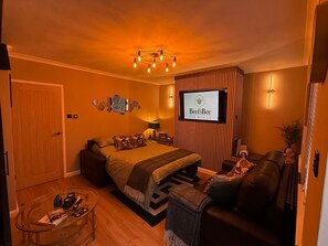 Quarto de hotel com cama, sofá, televisão exibindo 'Bee&Bee Stay with us' e luminária fixada na parede.