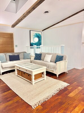 A living room with a white sofa, a glass coffee table, a wooden floor, and a ceiling with exposed beams. - The Beach Coop (Kill Devil Hills)