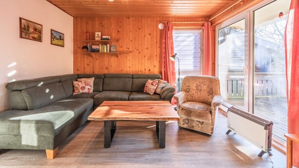 A cozy living room with a sectional sofa, a small round table, a patterned armchair, wooden walls, and a large window with curtains. - Familienfreundlicher Doppelbungalow mit Terrasse in Mirow (Mirow)