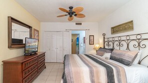 Ein Schlafzimmer mit Bett, Kommode, Deckenventilator, Fernseher und einer Tür zu einem anderen Raum.