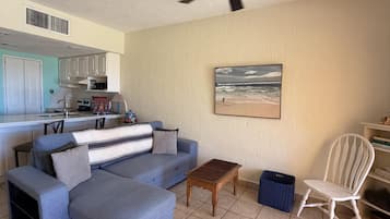 A living room with a sofa, a coffee table, a chair, a ceiling fan, a kitchen in the background, and a beach scene painting on the wall.