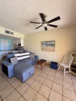 A living room with a sofa, a coffee table, a chair, a ceiling fan, a kitchen in the background, and a beach scene painting on the wall. - Princesa Resort D-105 - A Ground Floor Ocean View Condo (Puerto Peñasco)