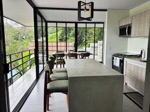 A modern kitchen with a large island, a dining area with chairs, and a view of greenery through large windows. - Modern Mountain Retreat: New 2-Bedroom Home in Beautiful Alto San Juan (alto san juan)
