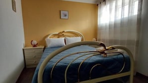 A bed with a metal frame and a blue bedspread, a bedside table with a lamp, and a window with sheer curtains. - Casa da Fonte (Peso da Regua)
