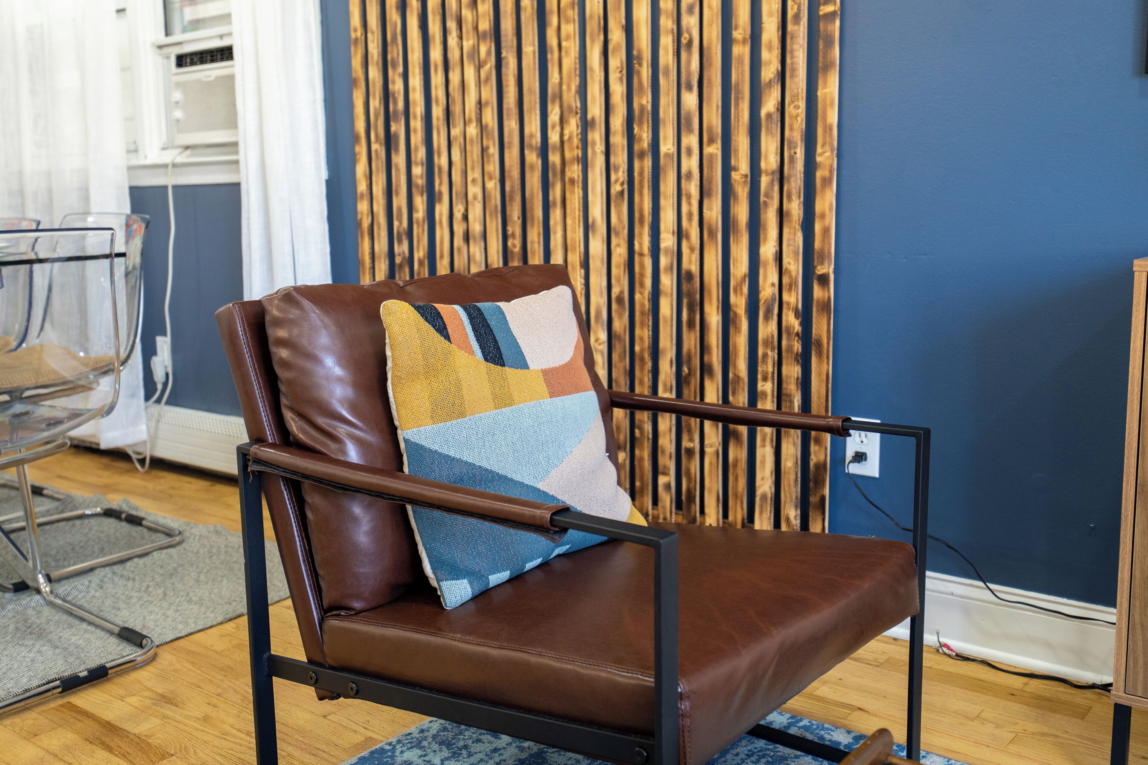 Un fauteuil en cuir marron agrémenté d’un coussin coloré, placé dans une pièce dont un mur est bleu et qui est dotée d’un fond en lambris de bois.