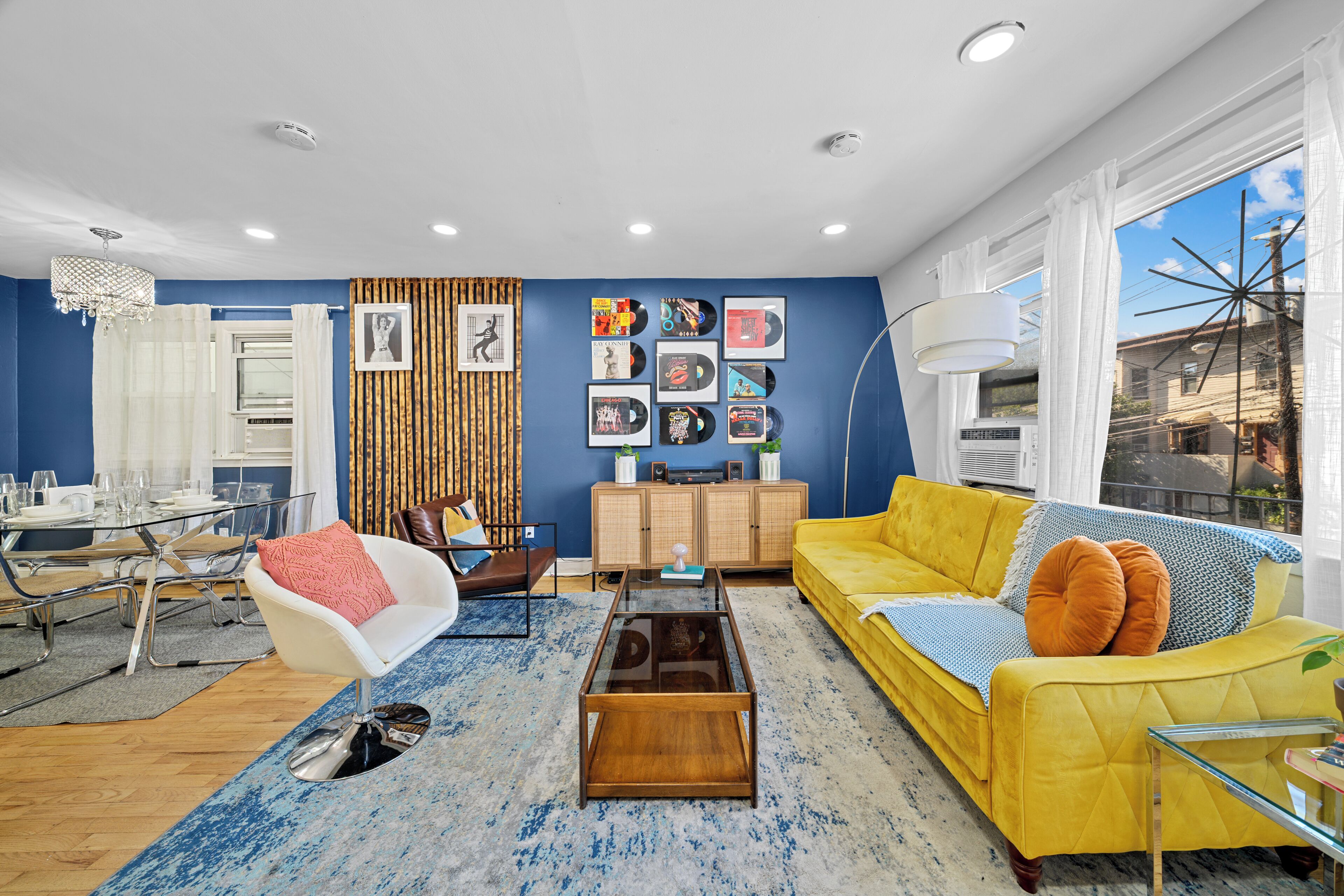 Una sala de estar moderna con un sofá amarillo, un sillón blanco, una mesa de centro de vidrio y un comedor con una mesa y sillas de vidrio.