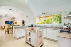 A modern kitchen with a central island, a dining area with chairs, and a large window with a view of greenery. - 4-Bedroom Villa with an Exclusive Private Pool, Sleeps 8 (playa del carmen)