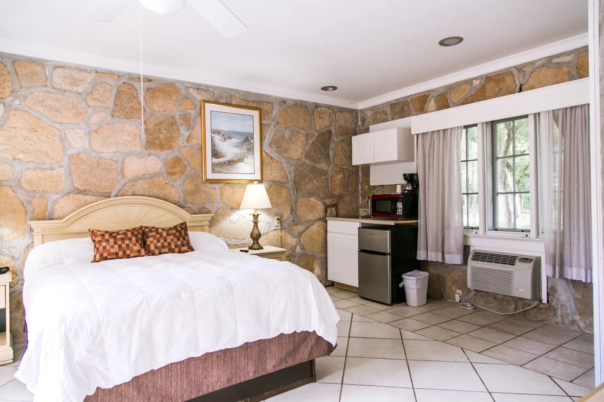 Quarto com parede de pedra, cama com almofada xadrez, mesa de cabeceira com abajur, cozinha compacta com micro-ondas e janela com cortinas.