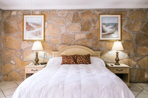 A bed with a white duvet, two nightstands with lamps, a stone wall, and two framed pictures. - Palm Coast Villas (Palm Coast)