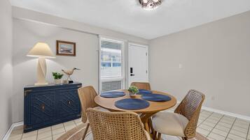 Un coin repas comprenant une table ronde, des chaises en osier, un buffet, une lampe et une porte avec des stores.