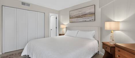 Quarto de hotel com uma cama grande, mesa de cabeceira, cômoda de madeira, um quadro na parede e um guarda-roupa.