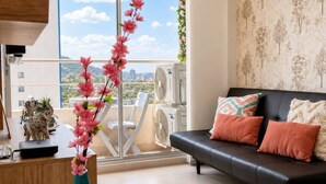 Ein modernes Wohnzimmer mit einem schwarzen Sofa, einem grünen Blumentopf mit rosa Blumen und einem Balkon mit Blick auf die Stadt.