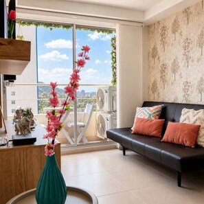 Ein modernes Wohnzimmer mit einem schwarzen Sofa, einem grünen Blumentopf mit rosa Blumen und einem Balkon mit Blick auf die Stadt.