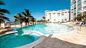 A swimming pool with a curved edge, surrounded by palm trees and a beach. There are lounge chairs and a building with multiple floors in the background. - Beach Apartment in Juan Dolio, Getaway Offer (Playa Juan Dolio)