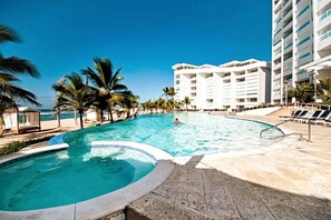 Ein Pool mit geschwungener Kante, umgeben von Palmen und einem Strand. Liegestühle und ein mehrstöckiges Gebäude sind im Hintergrund zu sehen.