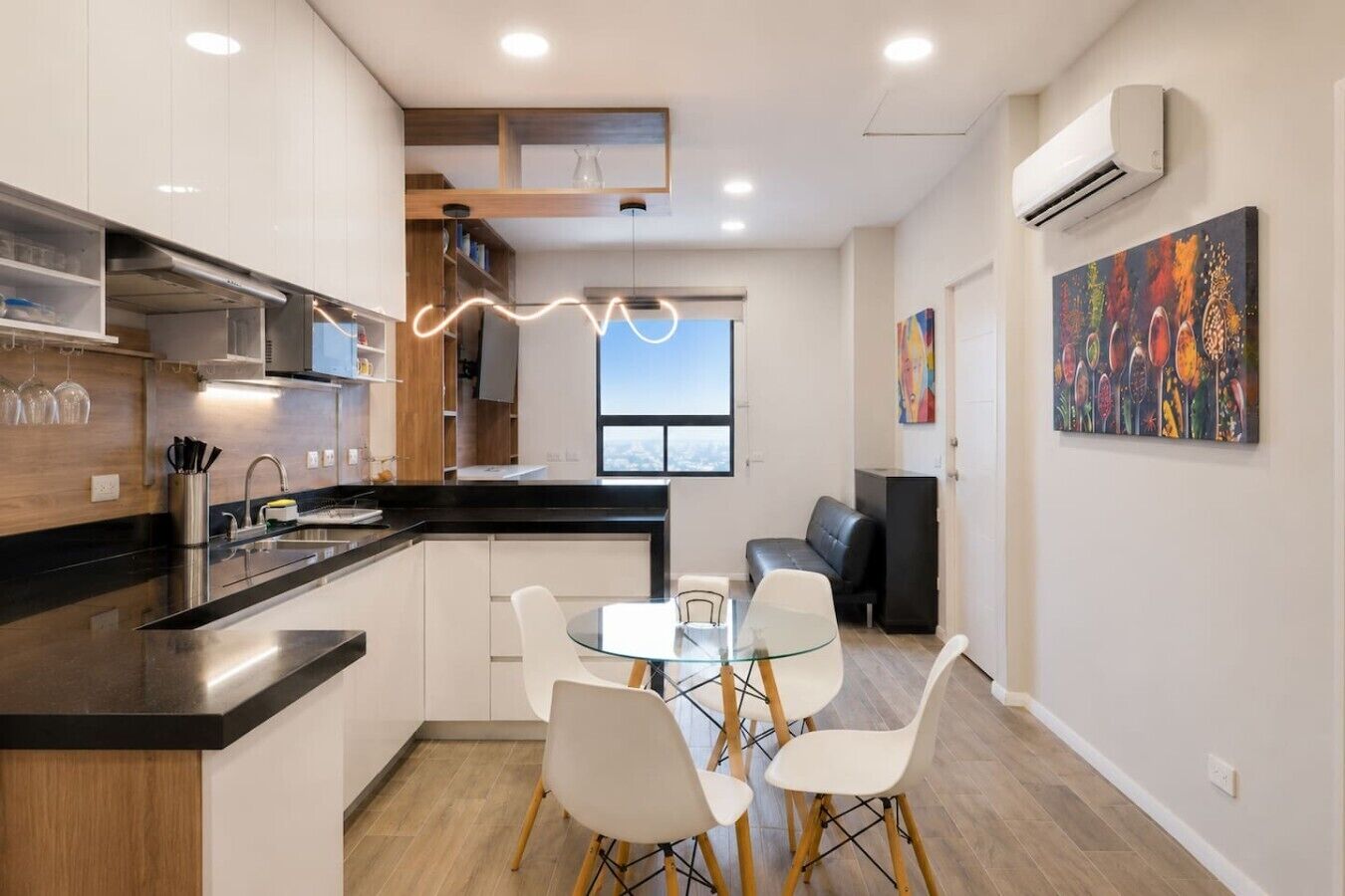 A modern kitchen with a dining area, a black countertop, white chairs, and a view of the city through a large window.