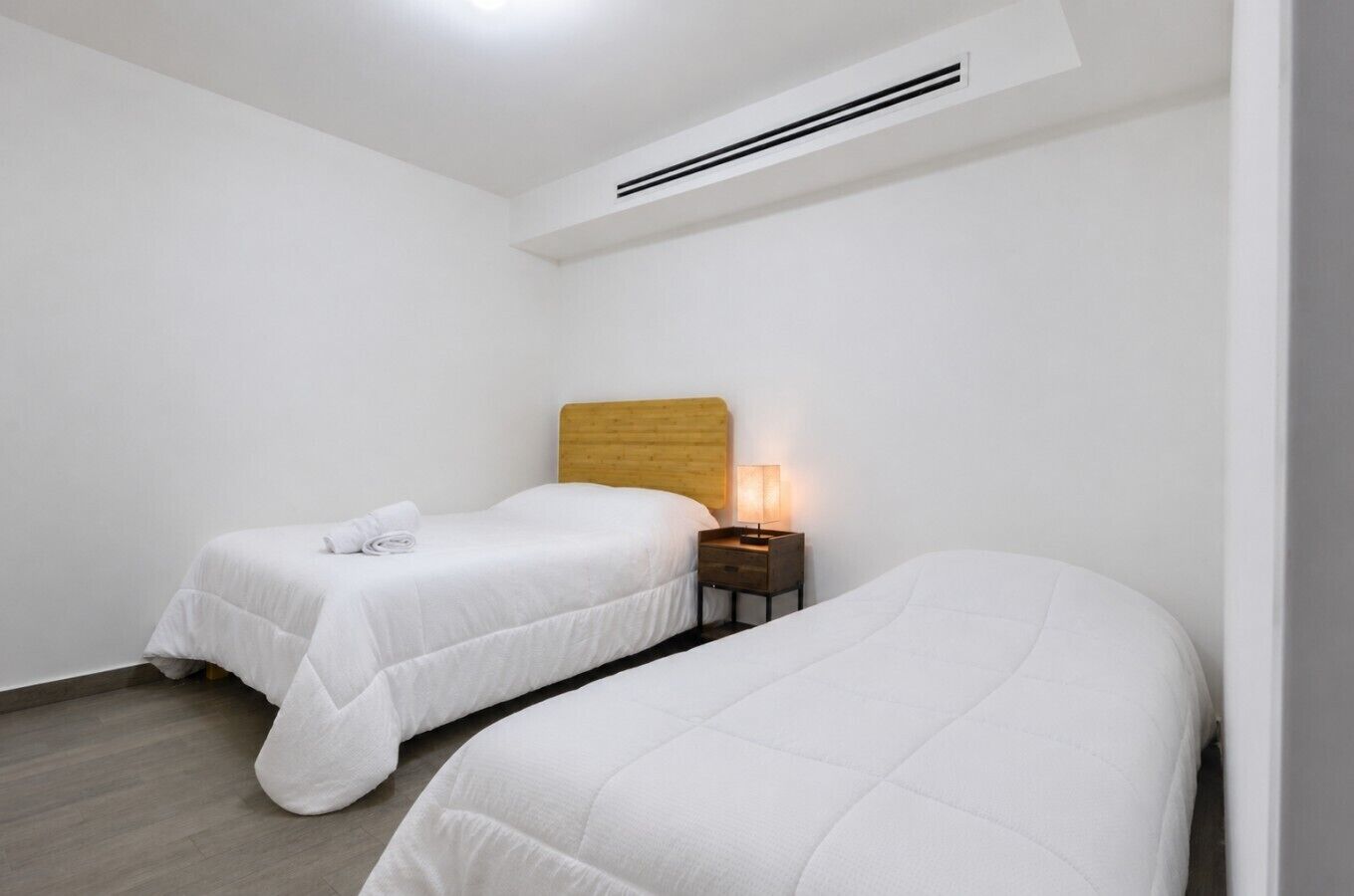 A room with two beds, a wooden headboard, a nightstand with a lamp, and a ceiling vent.