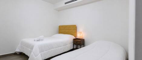 A room with two beds, a wooden headboard, a nightstand with a lamp, and a ceiling vent.