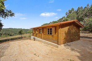 Una cabaña de madera pequeña con techo rojo, ubicada en suelo de tierra con vegetación escasa, rodeada por un paisaje montañoso.