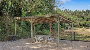 Un pavillon rustique en bois, doté d’une table et de bancs, entouré de gravier et d’arbres.