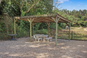 Ein rustikales Holzpavillon mit Tisch und Bänken, umgeben von Kies und Bäumen.