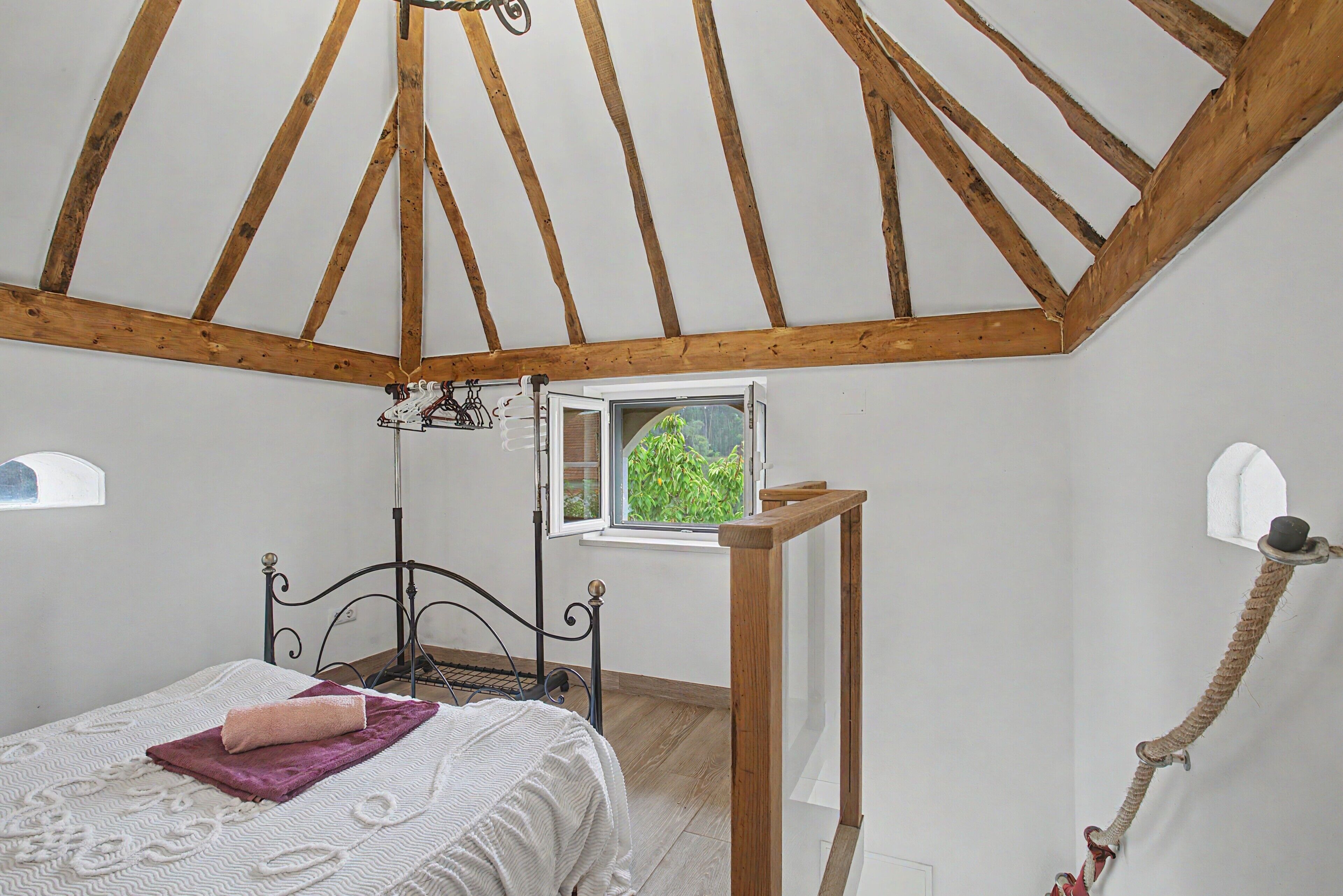A bedroom with a sloped ceiling, a bed with a headboard, a window with a view of greenery, and a rope ladder.