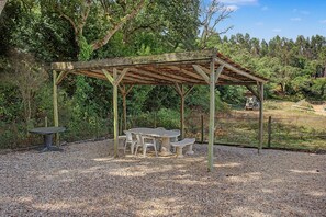 Ein rustikales Holzpavillon mit Tisch und Bänken, umgeben von Kies und Bäumen.