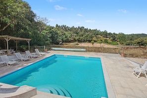 Ein rechteckiger Swimmingpool mit glasklar blauem Wasser, umgeben von einer gefliesten Liegefläche. Weiße Liegestühle und ein überdachter Sitzbereich mit Dach sind vorhanden.
