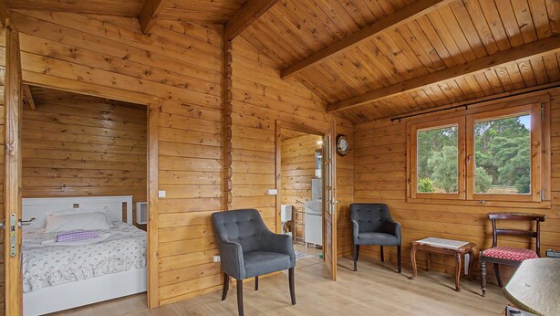 A wooden cabin interior with a bed, two armchairs, a small table, and a window with a view of trees.