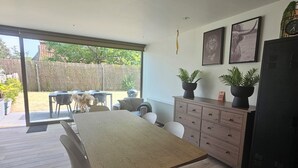 Une cuisine moderne avec un coin repas, comprenant une grande table, des chaises blanches, un réfrigérateur noir et une porte-fenêtre coulissante donnant sur un espace extérieur.