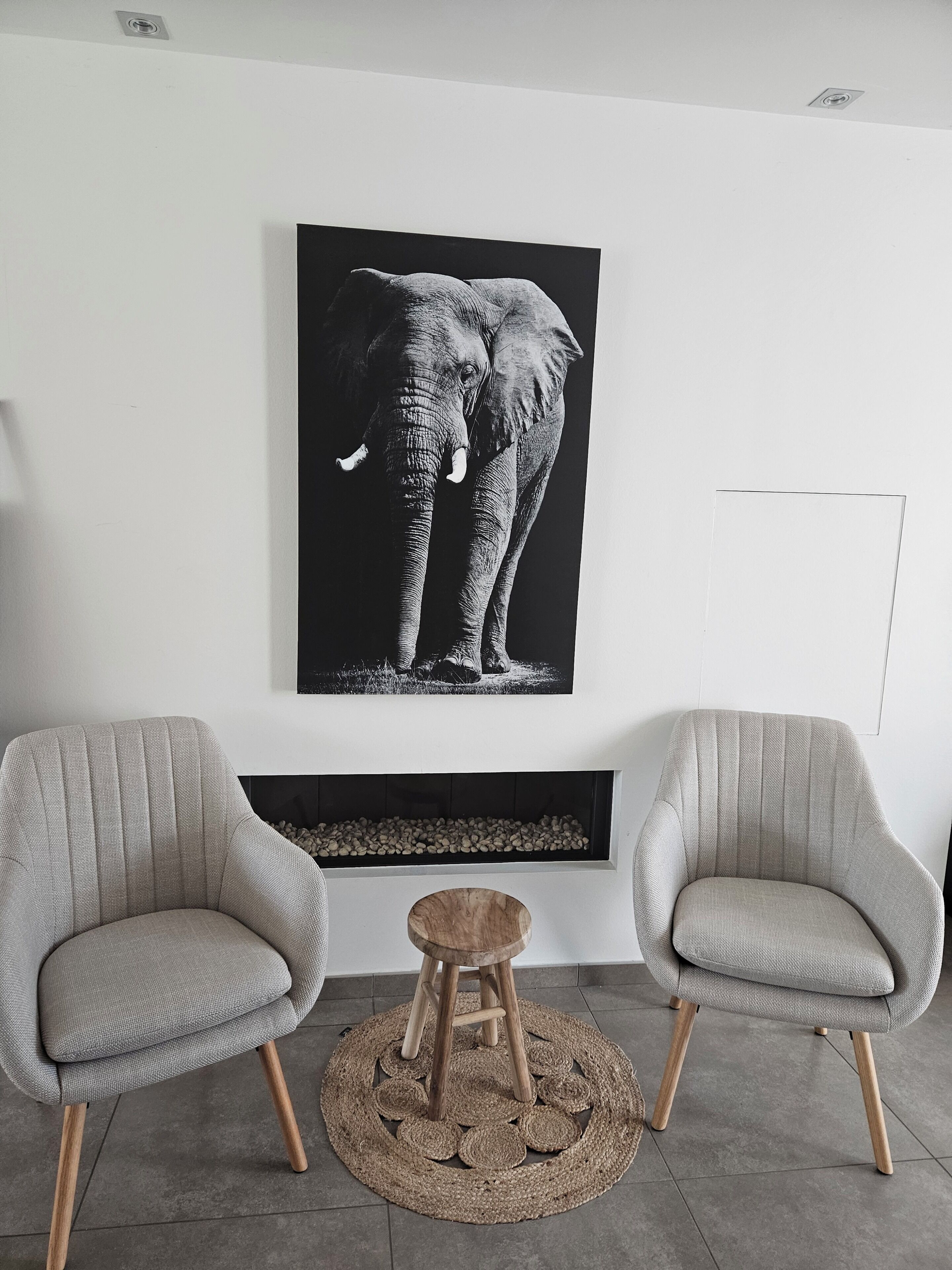 Un salon moderne avec deux fauteuils gris, un tabouret rond en bois, une cheminée et une grande image d’éléphant sur le mur.