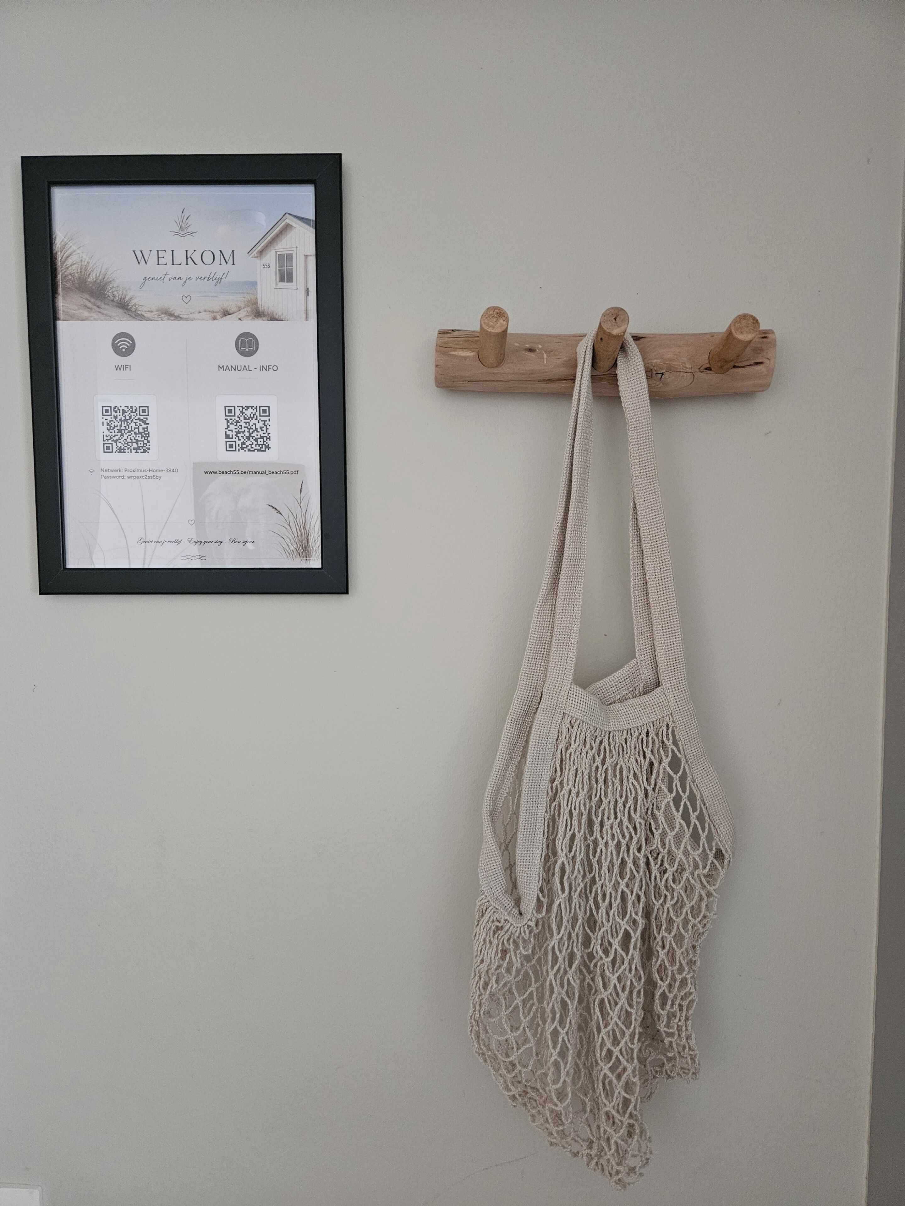 Un cartel de bienvenida en marco con códigos QR y una bolsa de macramé colgada en un perchero de madera.
