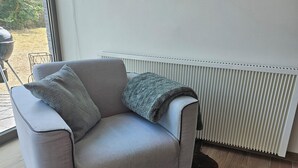 Un canapé gris clair agrémenté d’une couverture et de coussins, un tapis à poils longs de couleur marron foncé et un radiateur blanc dans une pièce dont le sol est recouvert de planches de bois.