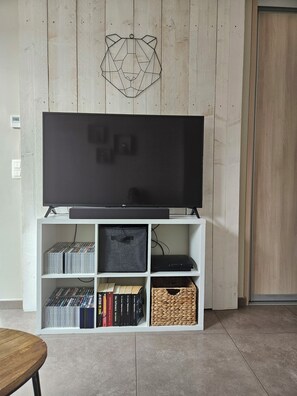 Un televisor de pantalla plana sobre un soporte blanco con repisas inferiores que contienen libros y una cesta tejida.
