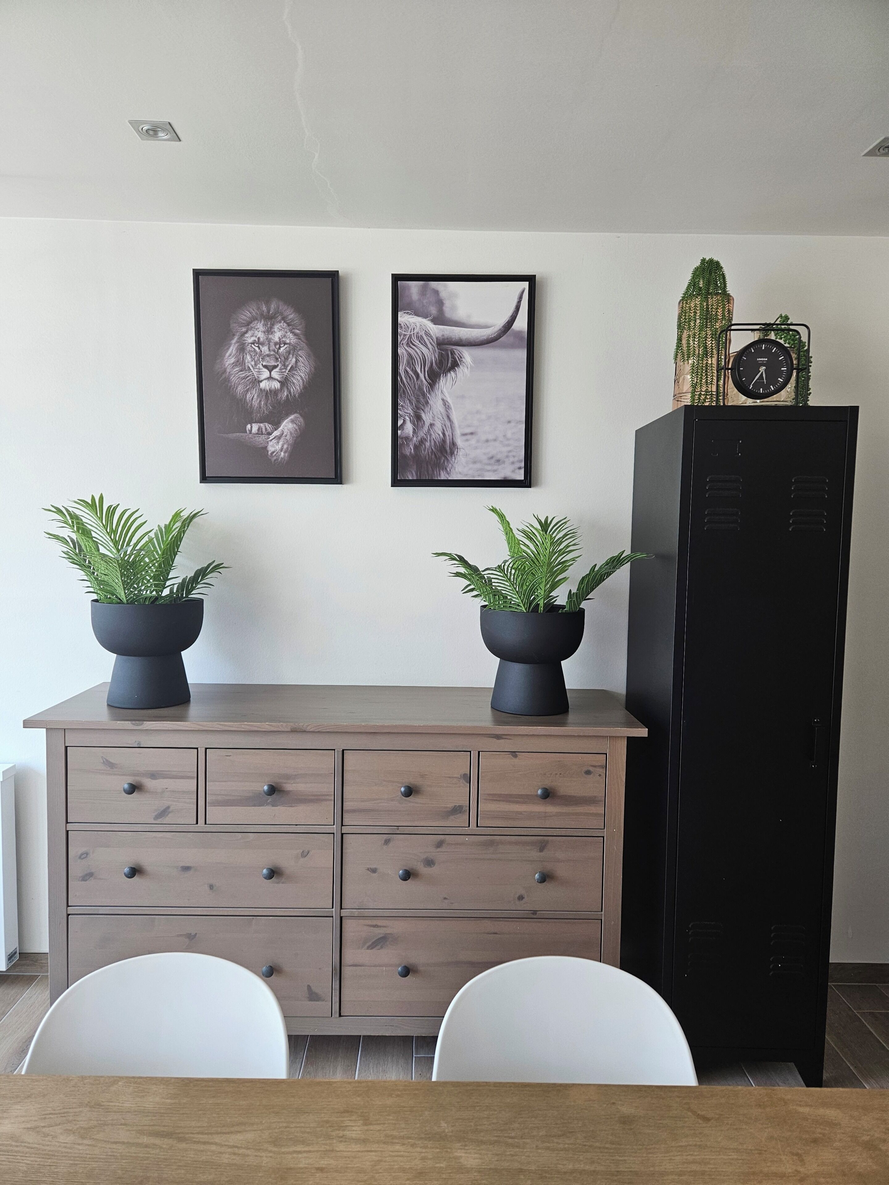 A dining area with a wooden table, two chairs, a cabinet with drawers, and two potted plants on the cabinet.