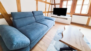 A room with a blue sofa, wooden beams, a TV on a white stand, and large windows. - Heinrich Apartment in a half-timbered house from 1596 – Jena’s Old Town (Jena)