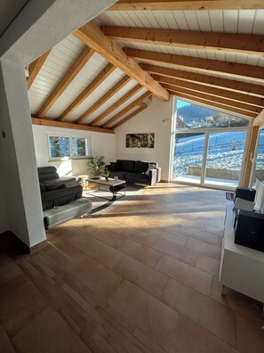 Un vaste espace de vie doté d’un plafond haut, de poutres en bois, d’un canapé, d’une table basse et offrant une vue sur un paysage enneigé grâce à de grandes fenêtres.