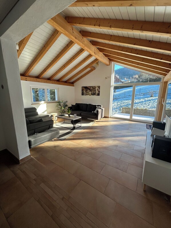 A spacious living area with a high ceiling, wooden beams, a sofa, a coffee table, and a view of a snowy landscape through large windows.
