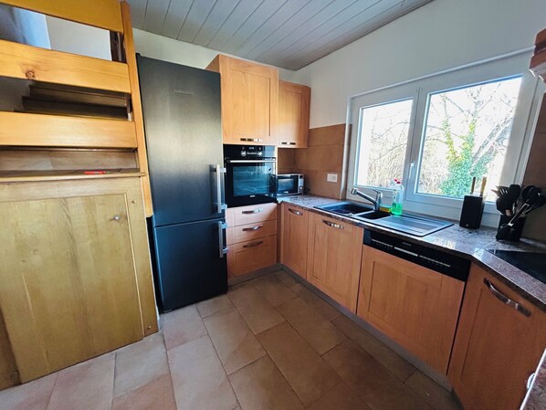Una cocina con armarios de madera, un refrigerador negro, un microondas y un ventanal con vistas a árboles.