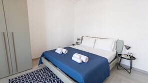 Un lit bien fait, avec une couvre-lit bleue et des oreillers blancs, une petite table de chevet avec une lampe, et un tapis à motifs sur le sol.