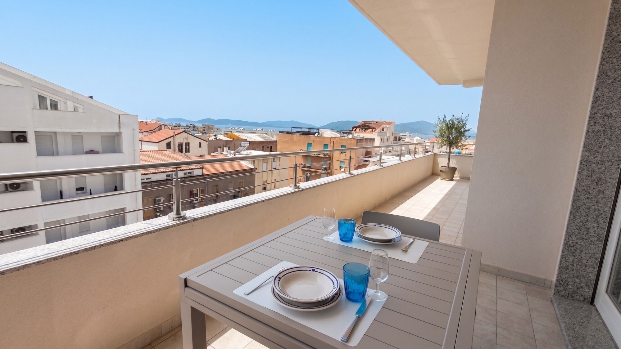 A balcony with a table set for two, overlooking a cityscape with buildings and mountains.