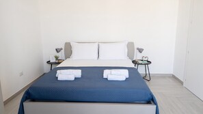 A neatly made bed with a blue bedspread and white pillows, two bedside tables with lamps, and a small potted plant on one table. - Sunset Panorama - Two Bedroom Apartment, Sleeps 4 (Alghero)