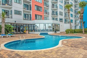 A swimming pool with a tiled deck, a building with balconies, and palm trees. - Palace 809 Ocean View Escape (Myrtle Beach)