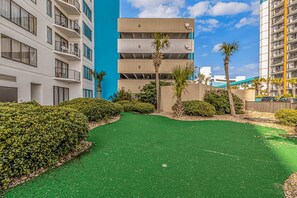 A courtyard with artificial green turf, palm trees, and a building with balconies. - Palace 809 Ocean View Escape (Myrtle Beach)