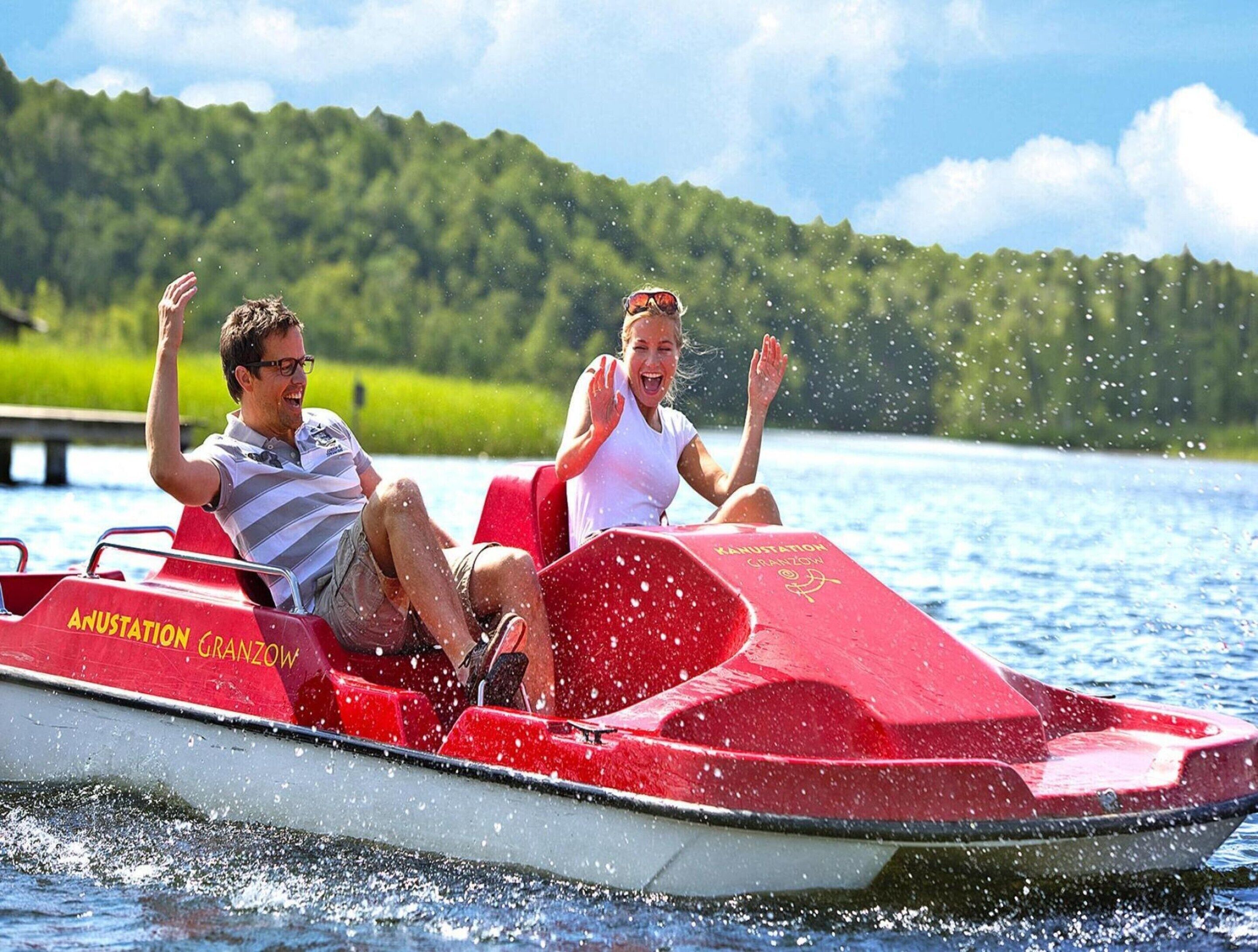 Two people on a red jet ski with "ANUSTATION GRANZOW" written on it, on a body of water with a forested hill in the background.