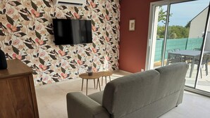 A living room with a grey sofa, a wooden cabinet, a small round table, and a TV mounted on the wall. - Les Parures d’Anthésim 20 : Cotentin – A/C & Green'Up EV Charger (Saint-Floxel)